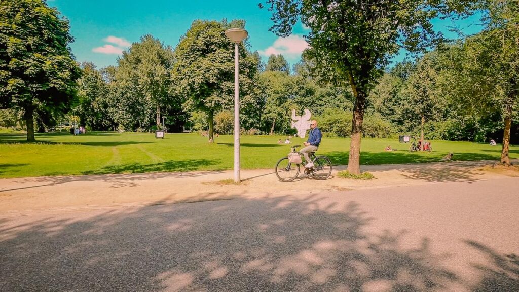 Cyclist enjoys a peaceful ride in Amsterdam's Vondelpark, with lush greenery and blue skies enhancing this perfect sunny day.