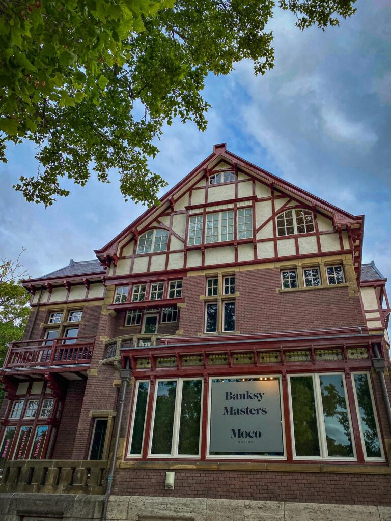 Moco Museum in Amsterdam featuring 'Banksy Masters' exhibition, vibrant red and white facade against green foliage and overcast sky.