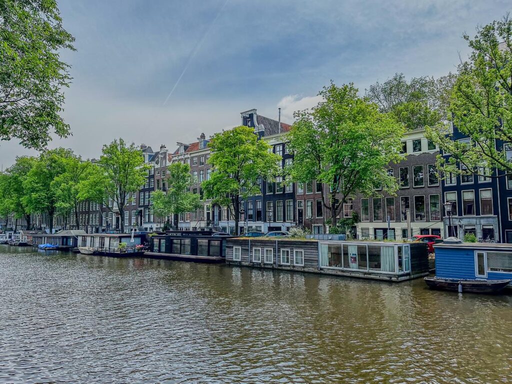 Historic Dutch houses and houseboats along the Singel canal in Amsterdam, embraced by lush foliage and a tranquil watery expanse, under a soft spring sky.